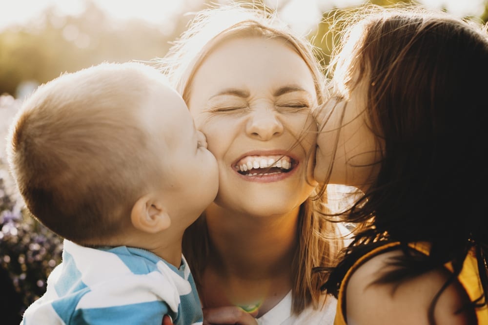 A mom with recently whitened teeth at West Richland Family Dental gets kissed on both cheeks by her young children